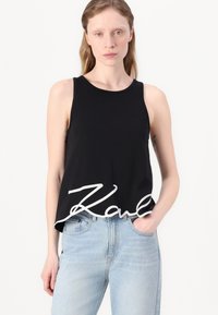 Svart ermeløs crop-top laget av myk stoff, med en stor hvit signaturtrykk "Karl" langs den nedre kanten. Matchet med lyseblå jeans.