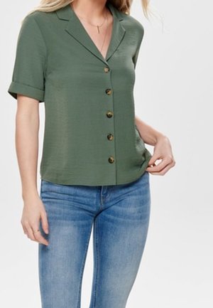 Femme portant un chemisier boutonné vert olive à manches courtes avec des poignets retroussés et un jean en denim bleu clair sur fond blanc.