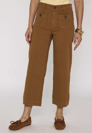 Pantalon marron large et court avec poches plaquées boutonnées à l'avant, porté avec des mocassins assortis et des bracelets en or.