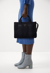 Černá látková taška na přenášení s dvojitými rukojeťmi a trojúhelníkovým kovovým logem. Hladká textura, obdélníkový tvar a detail zipové boční kapsy.