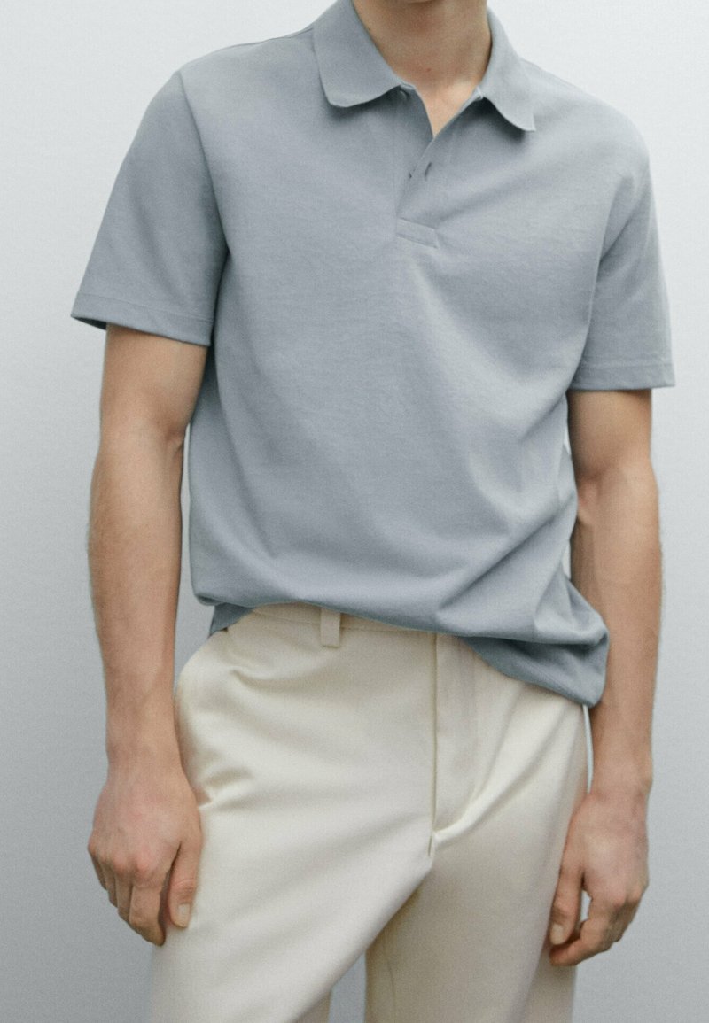 Lys grå polo shirt i blødt stof med klassisk krave og korte ærmer, parret med cremefarvede bukser. Simpelt design og pasform.