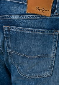 Jeans de mezclilla azul con un parche de cuero marrón claro que presenta la marca "Pepe Jeans London", bolsillo trasero con costuras visibles y un acabado texturizado.