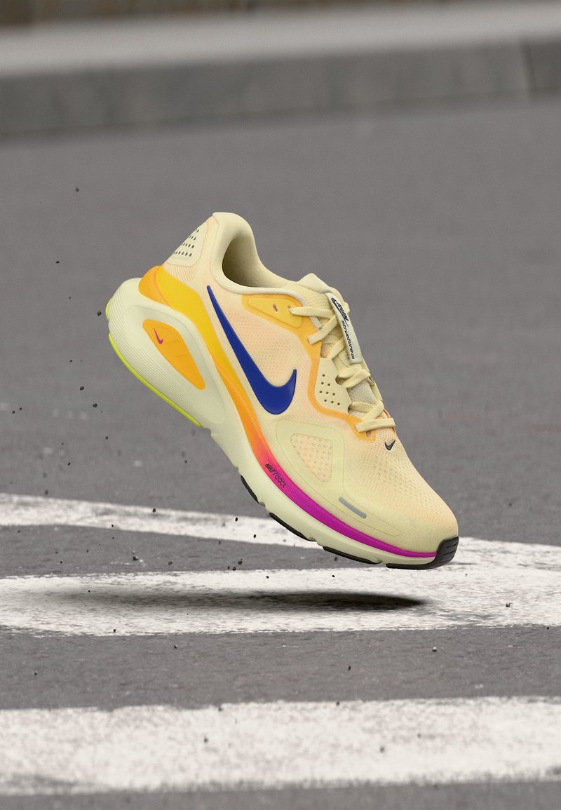 Chaussure de course Nike jaune clair avec le swoosh bleu, accents orange et rose sur la semelle, en suspension au-dessus d'une surface en asphalt gris avec des marquages blancs.