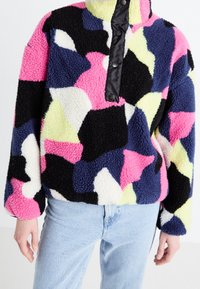 Fleece pullover med et flerfarvet abstrakt mønster i pink, blå, gul og sort, med en høj krave og trykknapdetalje.