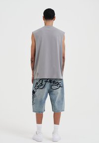 Camiseta gris sin mangas combinada con pantalones cortos de mezclilla sueltos y descoloridos que presentan diseños gráficos en negro y bordados, usados con zapatillas blancas.