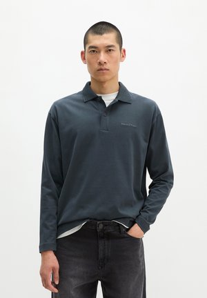 Homme aux cheveux courts portant un polo bleu foncé à manches longues et un jean noir, debout avec une main dans la poche sur fond blanc.