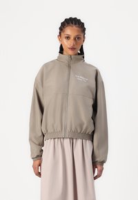 Lagani taupe windbreaker s visokim ovratnikom, elastičnom strukom i izvezenim logotipom. Duge rukave i glatku teksturu.