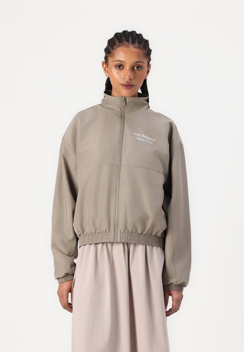 Lagani taupe windbreaker s visokim ovratnikom, elastičnom strukom i izvezenim logotipom. Duge rukave i glatku teksturu.