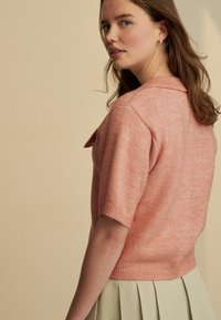 Kurzärmliger, hellrosa Strickpullover mit Kragen und Knopfdetail, getragen über einen plissierten, hellgrünen Rock, vor einem neutralen Hintergrund.
