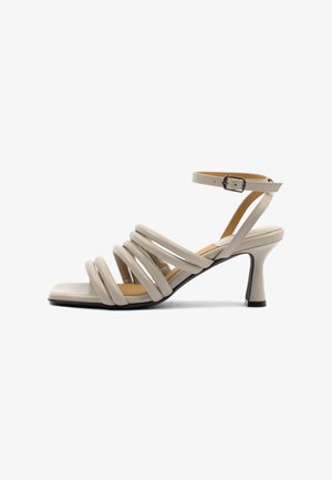 Sandalo beige da donna con tacco, cinturini incrociati imbottiti, cinturino alla caviglia con fibbia, punta quadrata e tacco medio svasato su sfondo bianco.