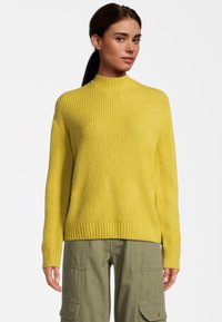 Gelber gerippter Strickpullover mit hohem Kragen und langen Ärmeln, kombiniert mit olivgrünen Cargo-Hosen. Die Textur wirkt weich und warm.