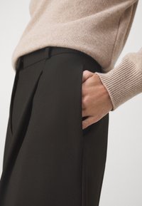 Main rentrée dans la poche d'un pantalon noir taille haute ajusté, porté avec un pull beige côtelé.