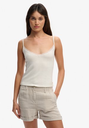 Femme portant un haut blanc sans manches en côtes avec bordure en dentelle et un short beige à ourlet roulé, debout avec une main dans la poche.