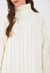 Suéter de cuello alto de punto trenzado en un suave blanco cremoso. Presenta un collar de canalé y un patrón texturizado en la tela para mayor detalle.