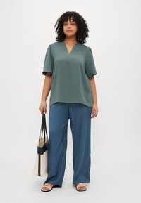 Τιρκουάζ μπλουζάκι κοντομάνικο με V λαιμόκοψη και υφή, συνδυασμένο με μπλε παντελόνι φαρδύ και κρατώντ ας μία κρεμ τσάντα.