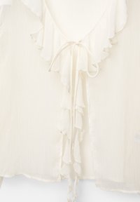 Blouse blanche avec un col froncé, dotée d'une fermeture par lien à l'avant. Fabriquée à partir d'un tissu léger et texturé avec des lignes verticales.