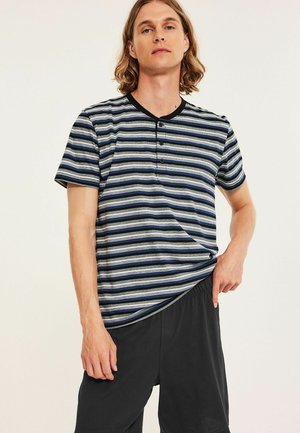 Jonge man met schouderlang haar, die een blauw, grijs en zwart gestreept, geknoopt t-shirt en zwarte korte broek draagt tegen een effen achtergrond.