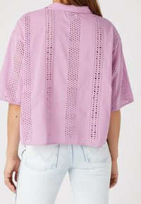 Osoba w liliowej bluzce z krótkim rękawem w ażurowy wzór i jasnoniebieskich jeansach z wysokim stanem, widziana od tyłu.