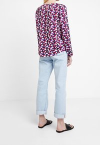 Blouse à manches longues avec un motif géométrique violet et rose, associée à un jean bleu clair retroussé aux chevilles et des sandales slides noires.