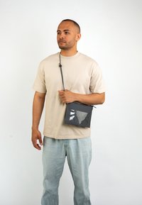 Bolso cruzado gris con una ventana transparente, cuenta con un cierre de cremallera y una correa ajustable. Se lleva de forma casual con una camiseta beige y jeans claros.