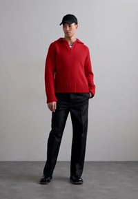 Red knitted sweater with a V-neck and ribbed hem, paired with black pinstriped trousers and a black cap, standing on a gray floor.
