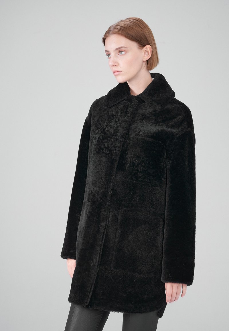 Schwarzer Kunstfellmantel mit Kragen, langen Ärmeln und einer Vordertasche. Der Stoff hat eine weiche, strukturierte Optik und einen lockeren Schnitt.