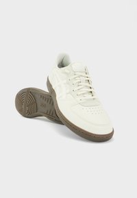 Paire de baskets en cuir blanc avec des accents beige et des semelles en caoutchouc marron, une chaussure positionnée droite et l’autre sur le côté.