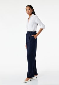 Chemise à col blanc et pantalon taille haute bleu marine à coupe ajustée, avec des plis et une texture lisse. Porté avec des chaussures à talons blanches.