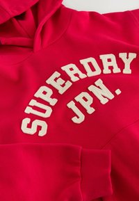 Červená mikina s kapucí a jemnou texturou, na přední straně s bílým vyšitým nápisem "SUPERDAY JPN." Obsahuje klokánkovou kapsu a žebrované manžety.
