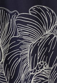 Motifs floraux blancs brodés avec des lignes courbes sur tissu bleu foncé, montrant une texture de couture détaillée et des pétales superposés.