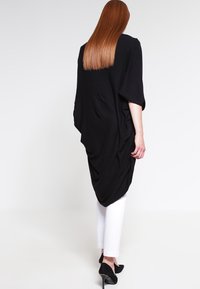 Svart överdimensionerad tunika med draperad design, långa ärmar och sidofickor, i kombination med vita byxor och svarta klackar. Matt textur.