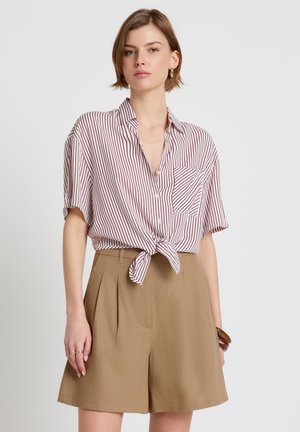 Donna che indossa una camicia a maniche corte, rossa e bianca a righe, abbottonata e legata in vita, con pantaloncini beige a vita alta e orecchini a cerchio.