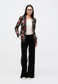 Blazer à motif floral en tissu foncé, à manches longues, avec un pantalon noir évasé. La tenue comprend un haut de couleur claire en dessous.