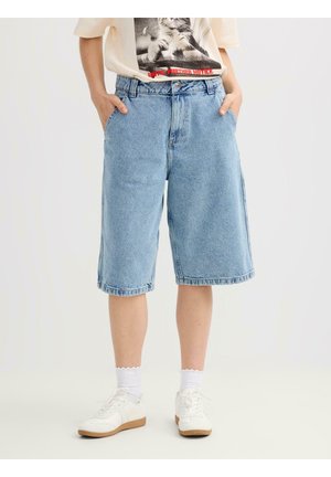 Persona que lleva puestos unos pantalones cortos de mezclilla azul claro hasta la rodilla, calcetines blancos, zapatillas blancas y una camiseta gráfica con las manos en los bolsillos.