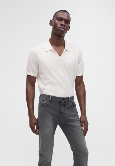 Homme portant un polo blanc à manches courtes et un jean gris, debout sur un fond blanc uni.