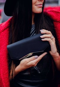 Black clutch bag with a sleek, angular design and metallic accent. Smooth texture, held by a person with a red coat and shimmering top.