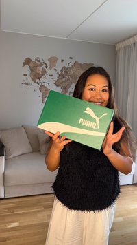 Zöld Puma cipősdoboz fehér díszítéssel és logóval, téglalap alakú, egy személy előtt tartva, aki texturált fekete felsőt és világos nadrágot visel.