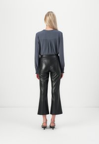 Graue Bluse mit langen Ärmeln und gerafftem Rücken, kombiniert mit hochgeschnittenen, schwarzen Faux-Leder Schlaghosen. Silbersandalen mit Riemen runden das Outfit ab.