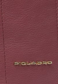 Superficie in pelle bordeaux con finitura testurizzata. Presenta il logo "Piquadro" in oro ben visibile. Cuciture ordinate lungo i bordi.