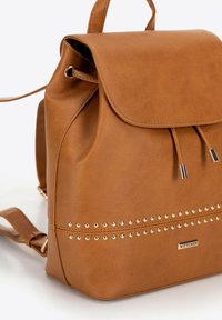 Hnědý batoh z umělé kůže s klopou, detaily na šňůrce a zlatými hroty podél spodní části. Má nastavitelné popruhy a logo značky.