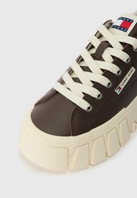 Smeđe kožne tenisice s kremasto bijelim gumastim potplatom, okruglim vrhom i kremastim vezicama. Ima oznaku s logotipom sa strane.