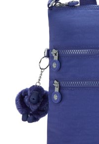 Lila nylon axelväska med två ytterfickor med dragkedja, prydd med en fluffig apcharm och en texturerad design.
