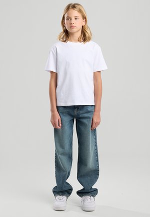 Enfant aux cheveux blonds mi-longs portant un t-shirt blanc uni, un jean bleu ample et des baskets blanches, debout sur un fond gris clair.