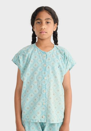 Dievča s tmavými vrkočmi nosí svetlomodrú blúzku na zapínanie a zladené nohavice s bielymi a žltými geometrickými kvetinovými vzormi.