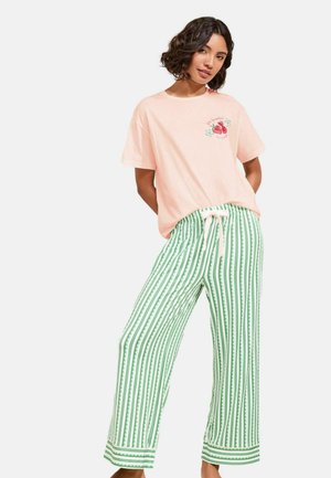 Femme portant un t-shirt rose clair avec un motif de fraise et un pantalon ample rayé vert et blanc à taille coulissée, debout les mains derrière le dos.