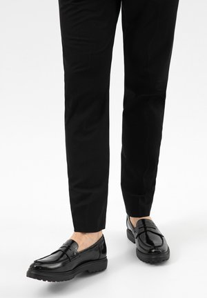Mocassini in pelle nera lucida indossati con pantaloni neri slim su sfondo bianco, piedi leggermente distanziati.