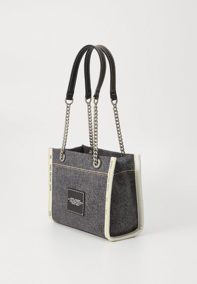 Sac fourre-tout en tissu gris foncé avec des poignées en cuir noir et enchaînement en argent, présentant des accents blancs et un patch de logo rectangulaire sur le devant.