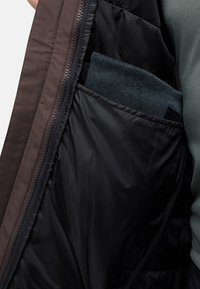 Giacca isolante marrone con una tasca interna nera, che mostra un articolo di maglieria blu scuro piegato all'interno. Texture liscia e opaca. Chiusura a zip.