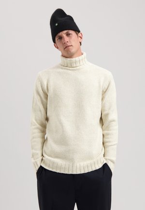 Kreemjas palus turtleneck särk ribidega nööpide ja servadega, koos musta beanie'ga, millel on ümmargune roheline aksent.