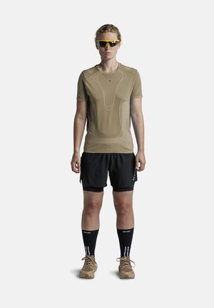 Atleet draagt een beige T-shirt met korte mouwen, zwarte korte broek, zwarte kousen tot over de kuiten, beige trailschoenen en gele sportzonnebril, staand met het gezicht naar voren.